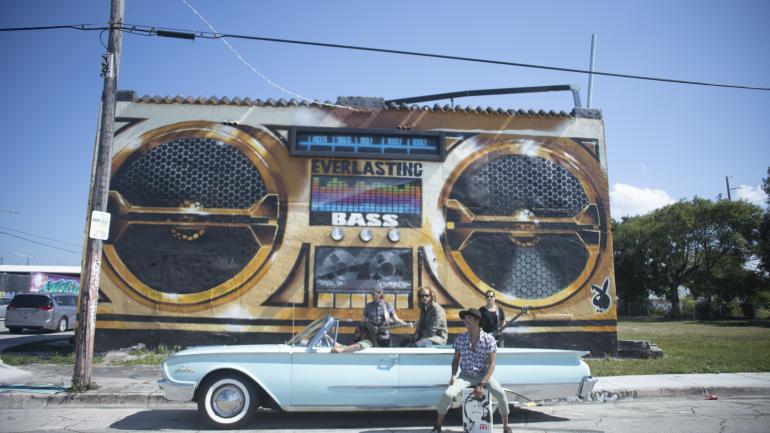 A banda posando com um mural de Miami com tema musical